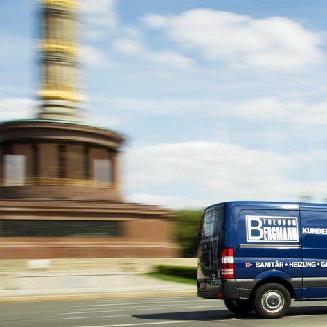 Der Lieferwagen von Bergmann Kundendienst ist in Bewegung, während die goldene Statue im Hintergrund verschwommen erscheint.