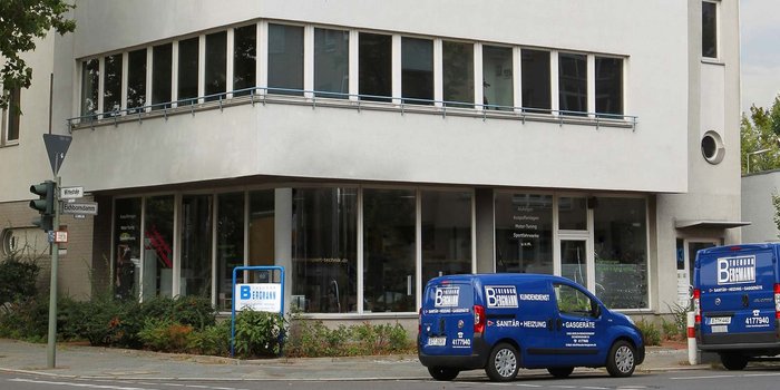 Das Gebäude an der Ecke hat eine klare, moderne Architektur und wird von zwei blauen Fahrzeugen des Unternehmens Bergmann flankiert.