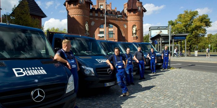 Die Gruppe von Arbeitern posiert vor ihren Fahrzeugen, während im Hintergrund ein markantes, rotes Gebäude sichtbar ist.