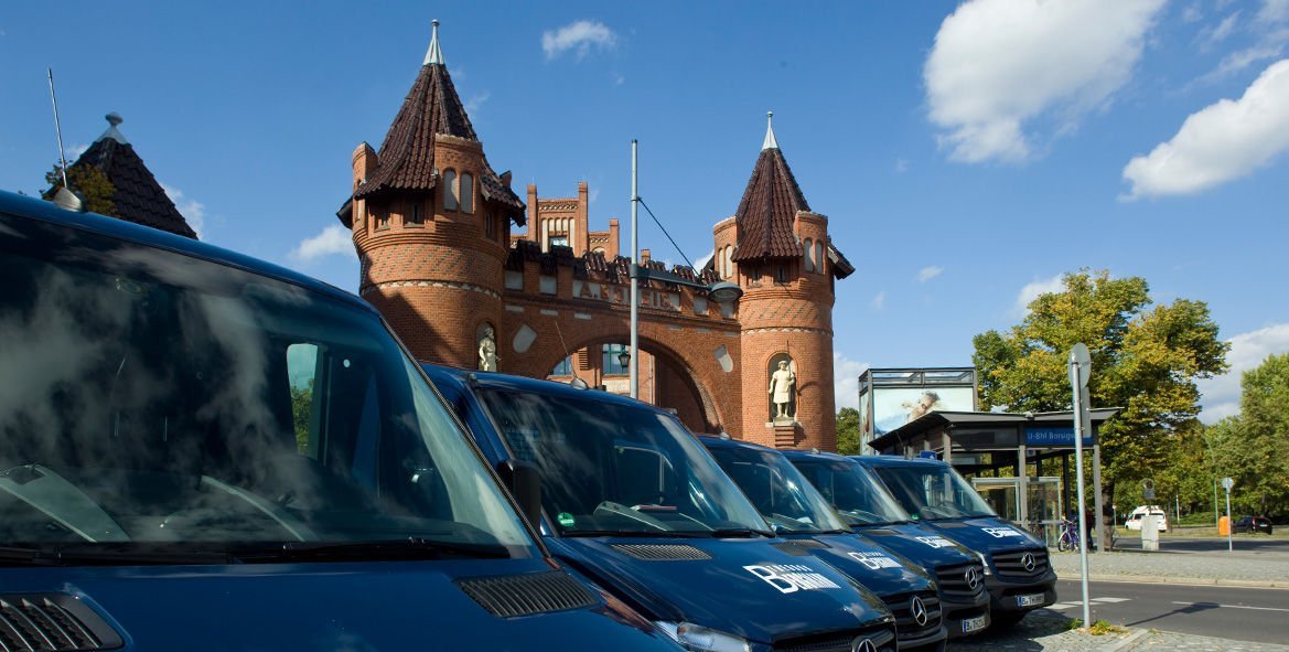 Die Szene zeigt mehrere Lieferwagen vor einem markanten, roten Tor mit Türmen und einer Statue, umgeben von Bäumen.